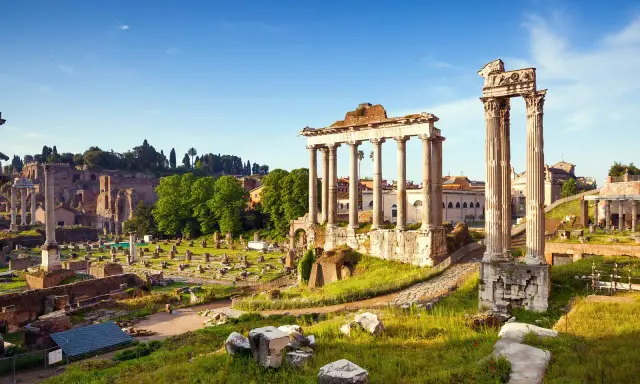 Antiche rovine del Foro Romano illuminate dal sole, con colonne imponenti e resti di templi sotto un cielo azzurro.
