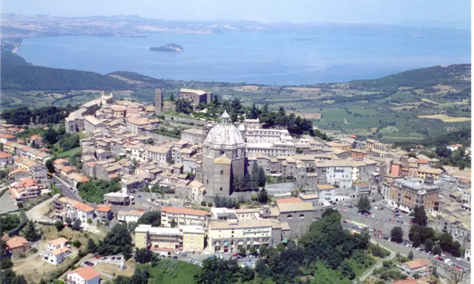 Vista aerea di un borgo italiano con cupola imponente, circondato da case con tetti rossi e affacciato su un lago. Est est est.