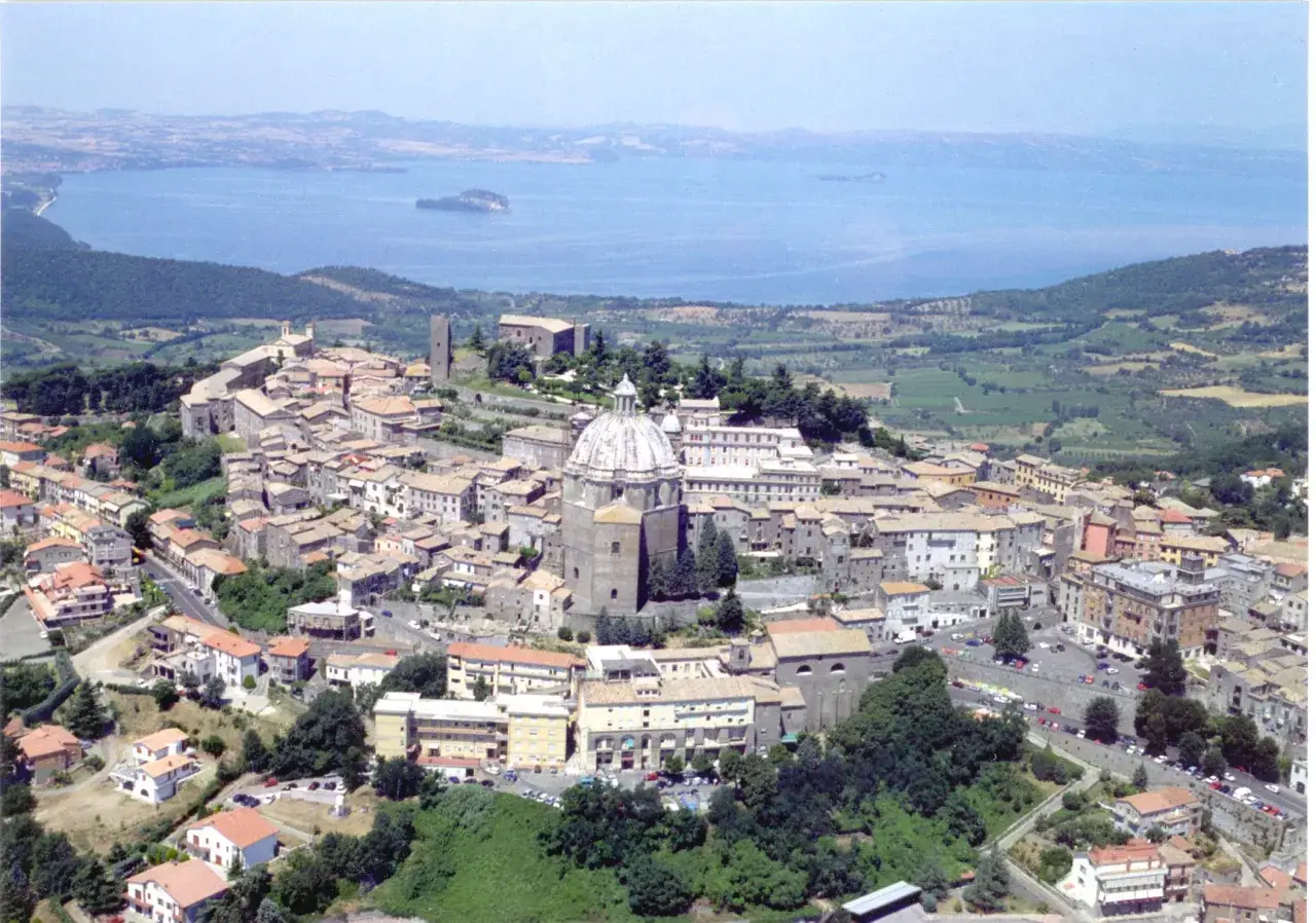 Vista aerea di un borgo italiano con cupola imponente, circondato da case con tetti rossi e affacciato su un lago. Est est est.