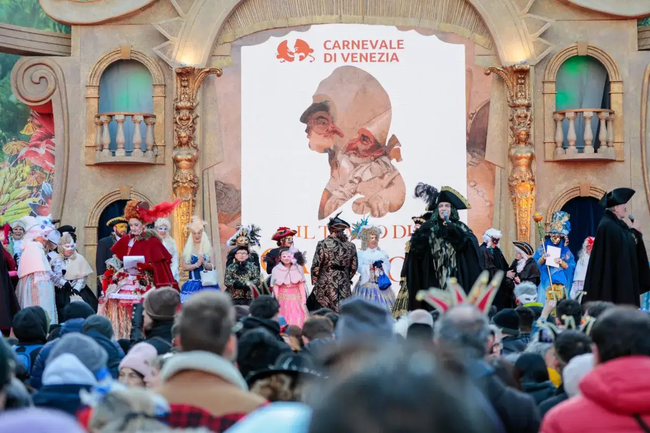 Persone in costume sfilano sul palco per il Carnevale di Venezia. Un grande schermo mostra il logo
