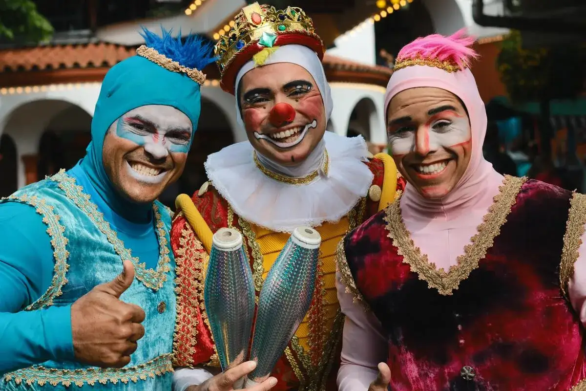 Tre artisti con maschere di carnevale siciliane, truccati da clown, sorridono e tengono in mano clave scintillanti.