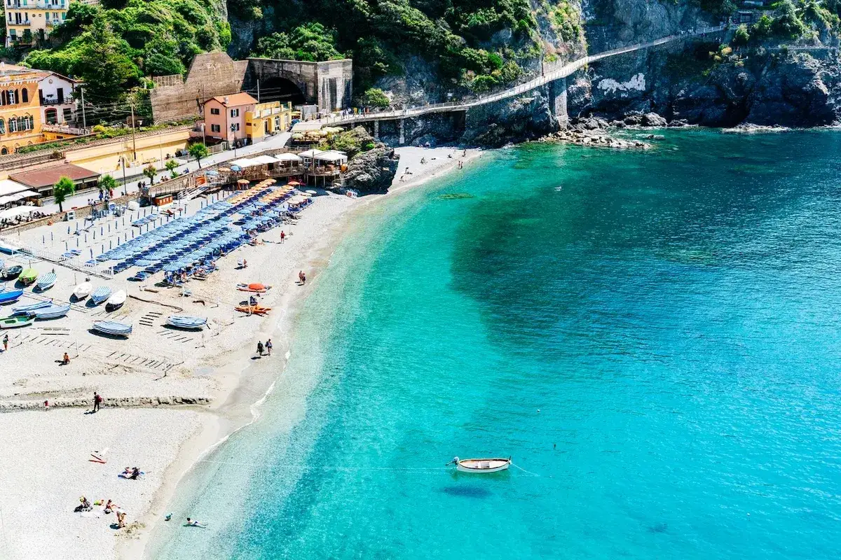 Un mare bello in Liguria, con una spiaggia attrezzata, barche sulla sabbia e un sentiero panoramico a picco sull'acqua cristallina.