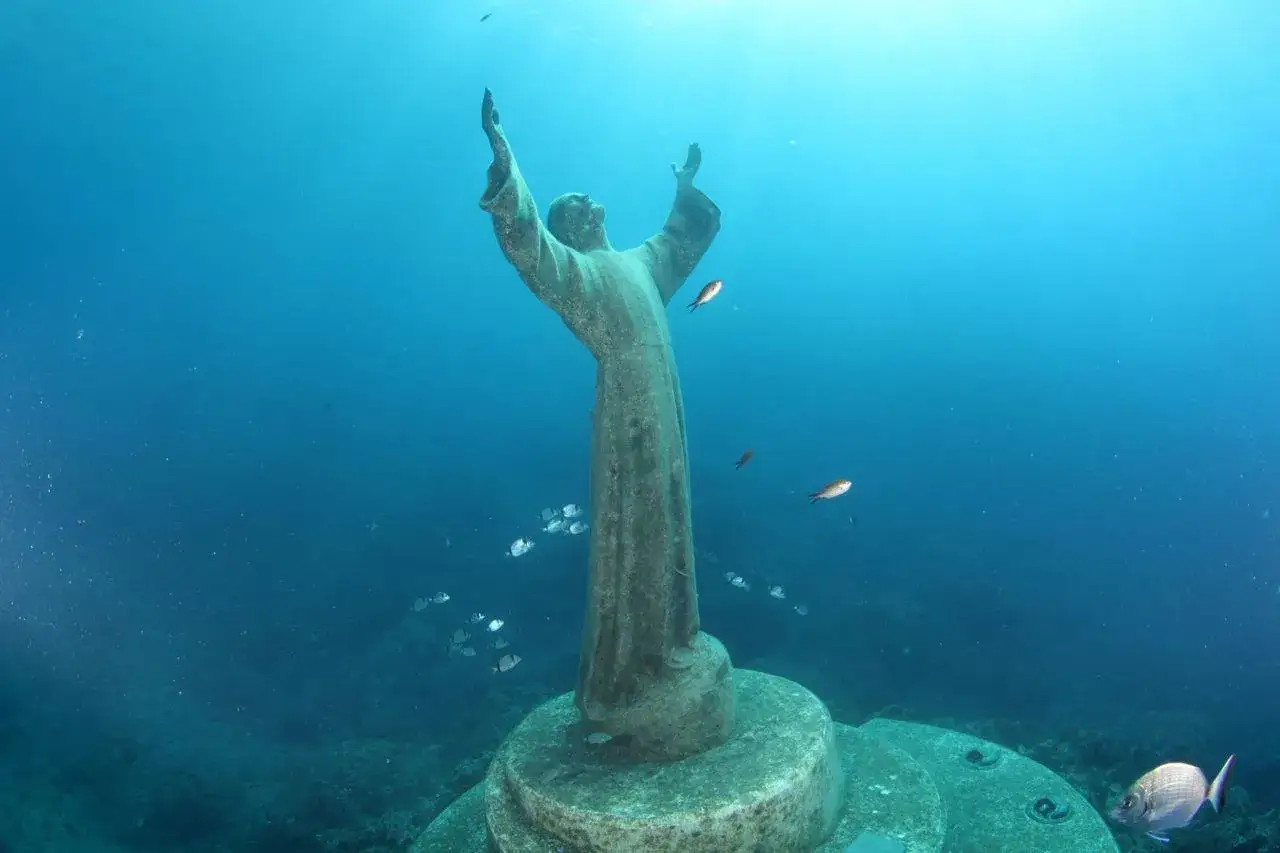Il Cristo degli Abissi, con le braccia alzate, veglia sui fondali marini, circondato da pesci che nuotano sereni nell'azzurro profondo.