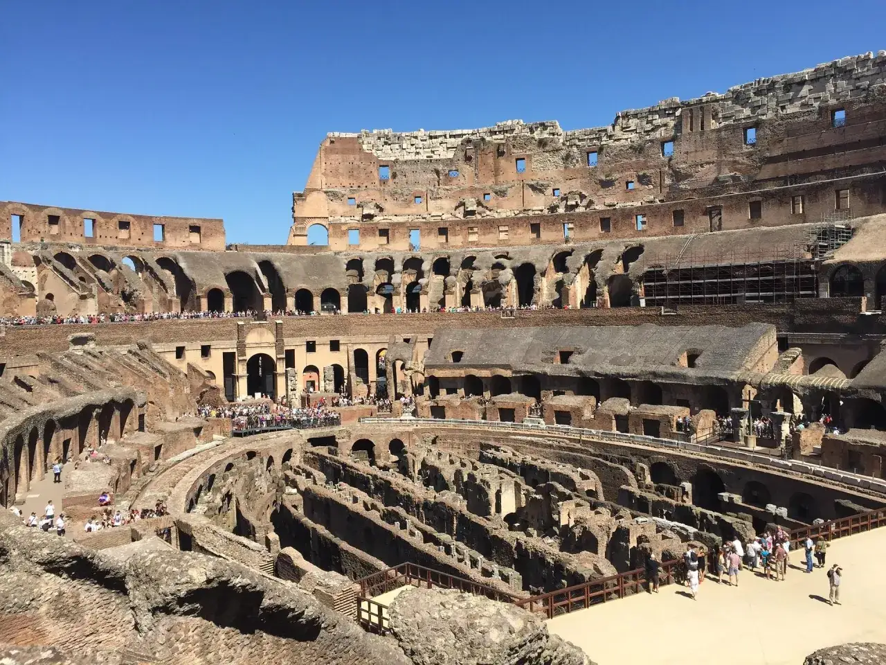 Interno del Colosseo con turisti. Le arcate e le strutture interne sono visibili, mostrando perché il Colosseo è bucato, con aperture che rivelano il cielo azzurro.