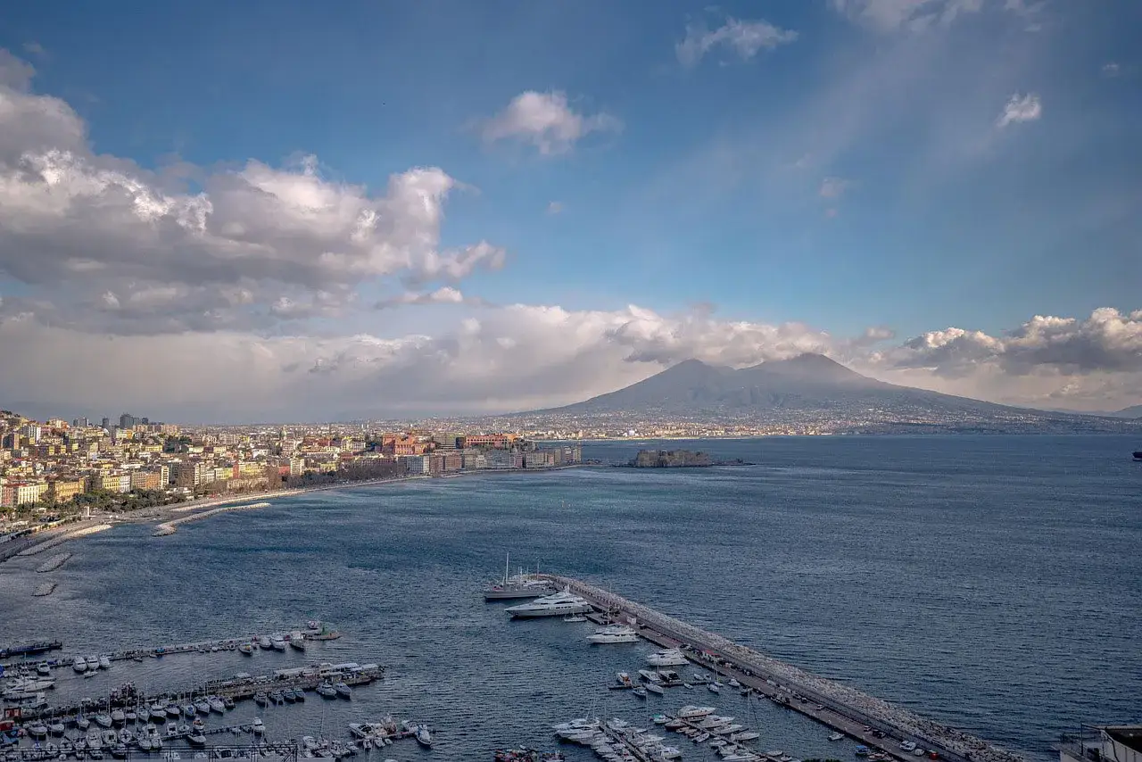 Vista panoramica di Napoli città con il Vesuvio sullo sfondo, porticciolo pieno di barche e il mare blu.