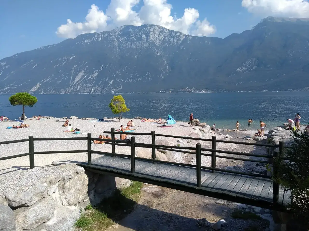 Spiaggia con gente che prende il sole e si bagna sul Lago di Garda, con montagne sullo sfondo. Un ponticello in legno attraversa un piccolo ruscello.