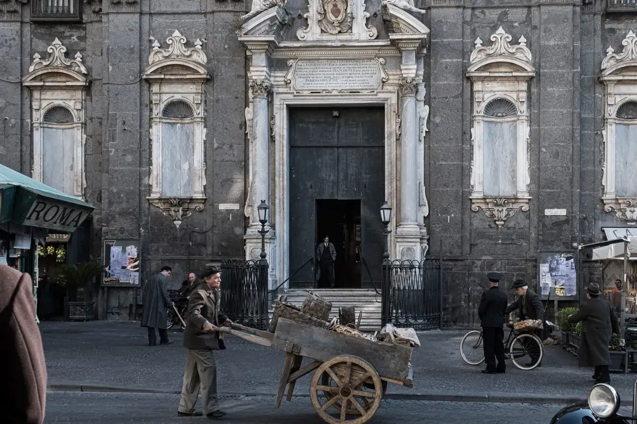 Uomo spinge un carretto per strada, un'espressione di fatica che riflette il senso del dolore. Architettura imponente sullo sfondo.