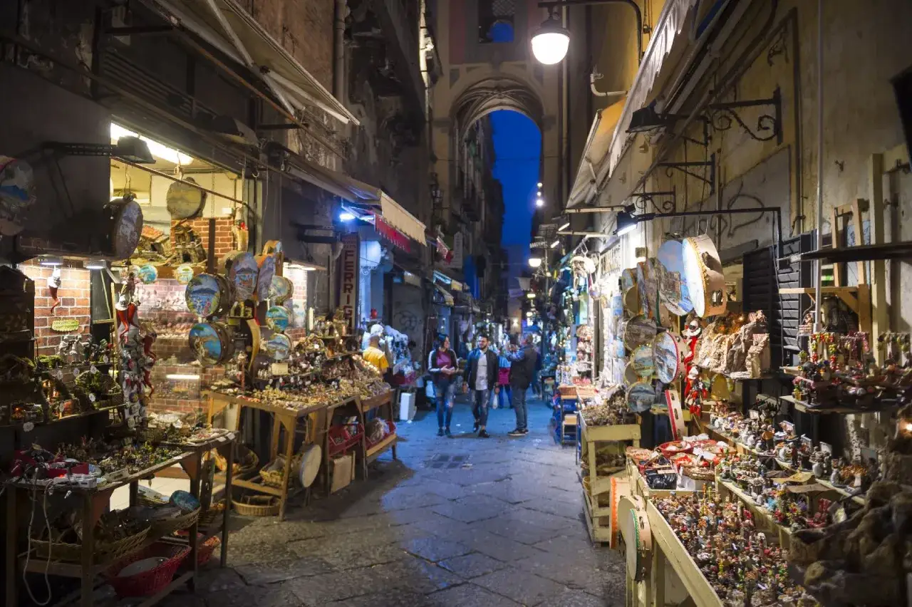 La via di San Gregorio Armeno, Napoli, illuminata di sera, con botteghe piene di statuine e oggetti artigianali.