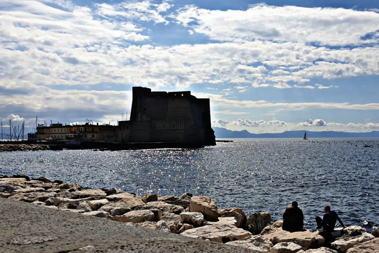 Castel dell'Ovo domina il golfo, un'icona di Napoli città. Due persone osservano il mare scintillante, con una barca a vela in lontananza.