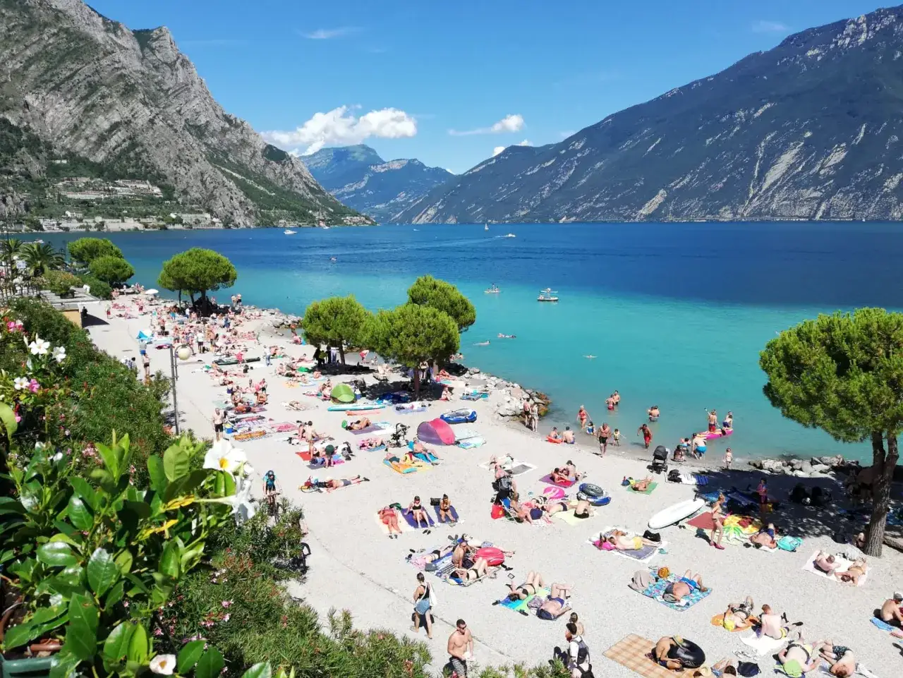 Spiaggia di Limone sul Garda affollata di bagnanti sotto le montagne. Acqua cristallina e sole splendente.