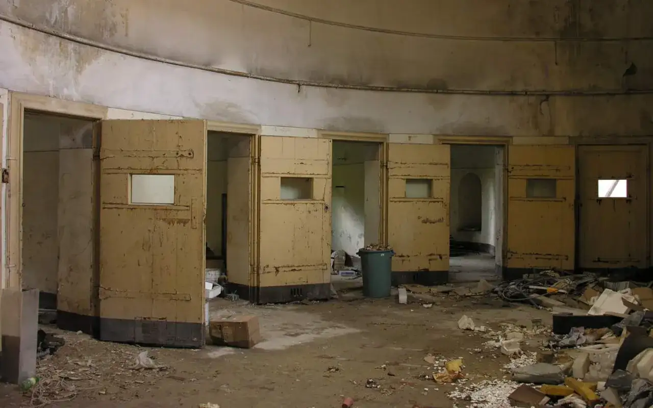 La sala delle agitate, un luogo abbandonato con porte aperte su celle vuote e detriti sparsi ovunque.