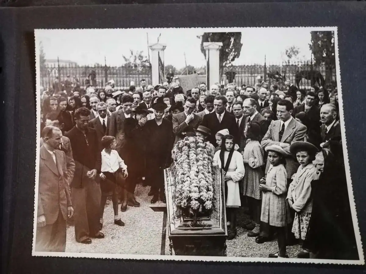 Una folla partecipa a un funerale, con un feretro fiorito al centro. Questa foto post mortem cattura un momento di lutto collettivo.