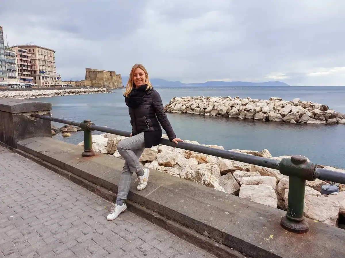 Un week end a Napoli: donna sorridente appoggiata a un parapetto con Castel dell'Ovo e il mare sullo sfondo.