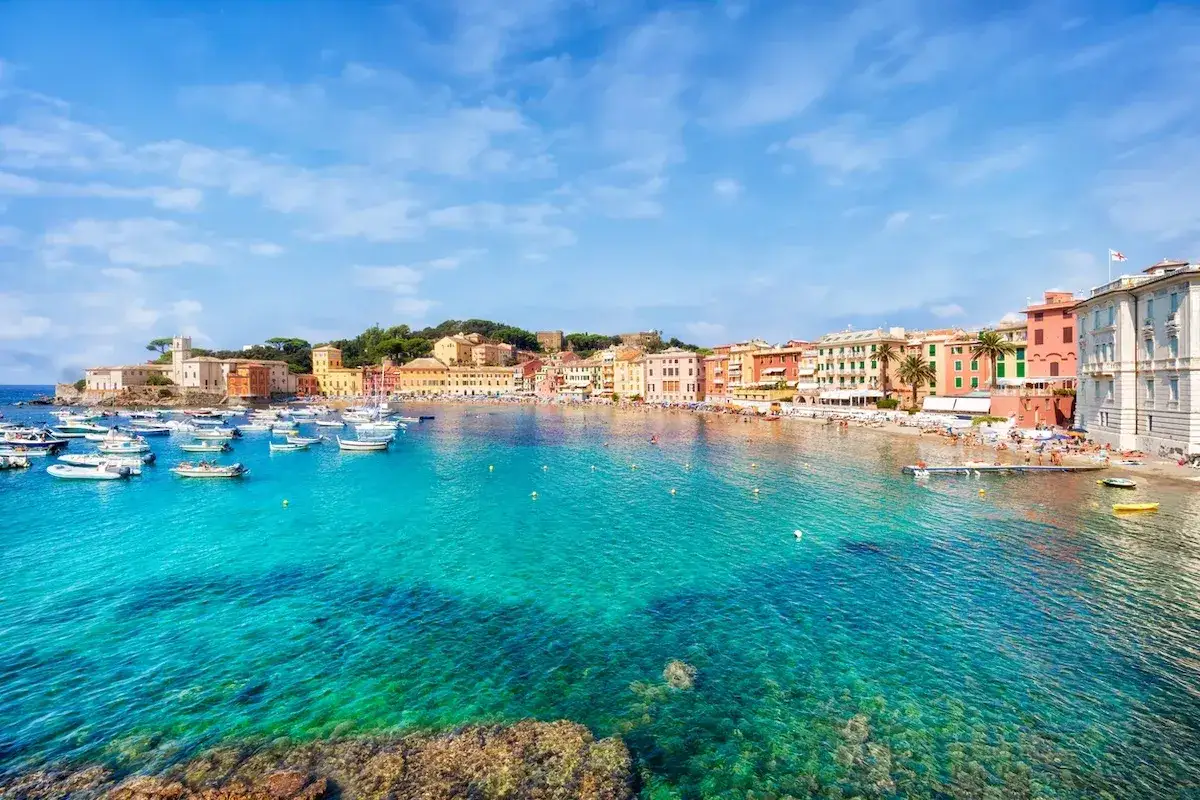 Un mare bello in Liguria, con acque cristalline e un borgo colorato affacciato sulla spiaggia. Barche a vela e piccole imbarcazioni ormeggiate.