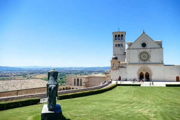 Statua equestre domina il prato verde di Assisi. Cosa vedere: la Basilica di San Francesco, con la sua imponente facciata e campanile, si staglia contro il cielo azzurro.