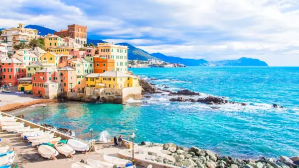 Case colorate a picco sul mare turchese, barche ormeggiate e un promontorio roccioso: un angolo di Genova che ricorda le sue spiagge più belle.