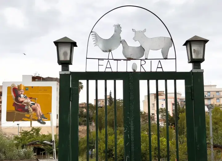 Cancello verde con insegna "FATTORIA" e sagome di animali. A sinistra, murale di un uomo su trono. Danisinni, Palermo.